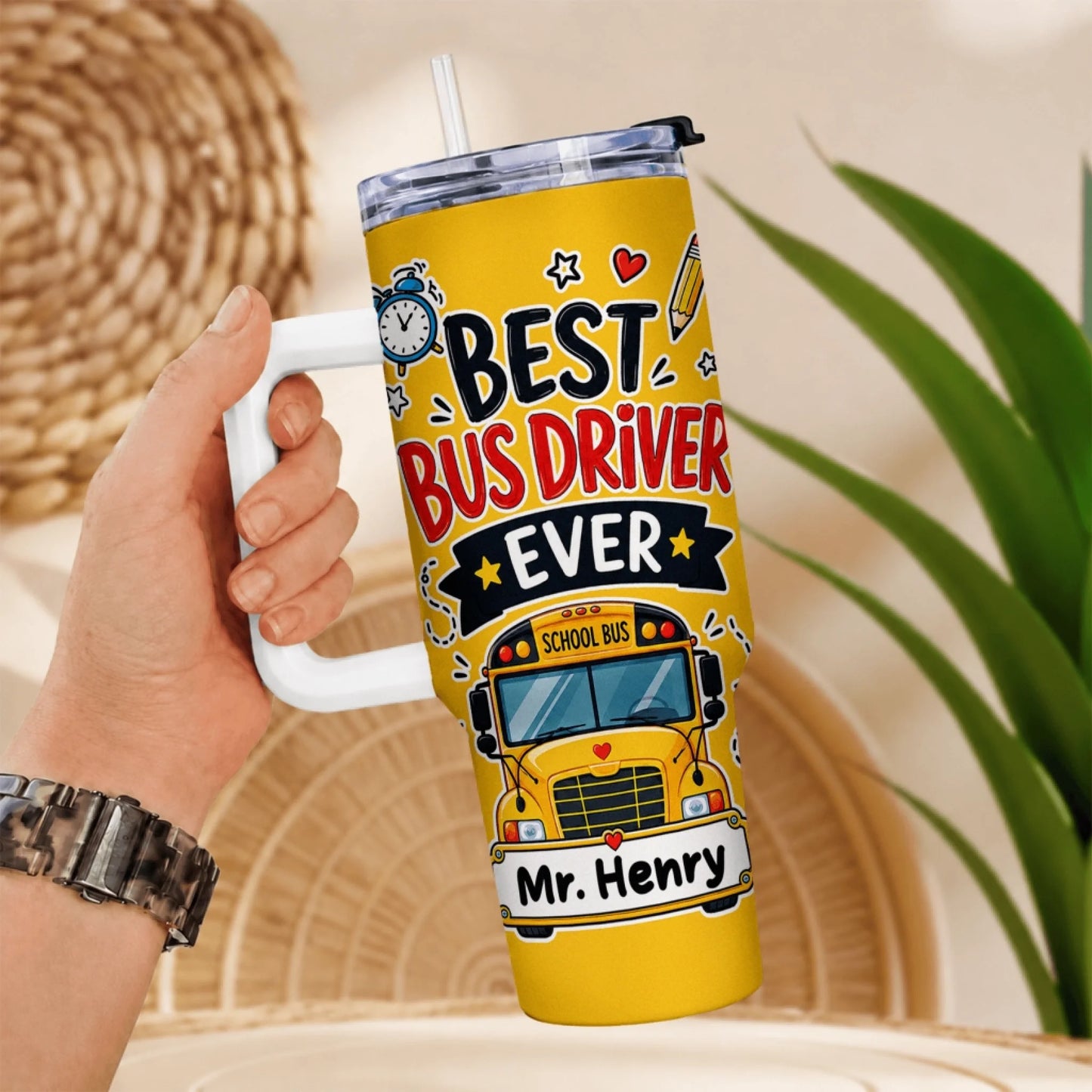 A hand holds a yellow tumbler with a white handle and straw, featuring a 'BEST BUS DRIVER EVER' design.