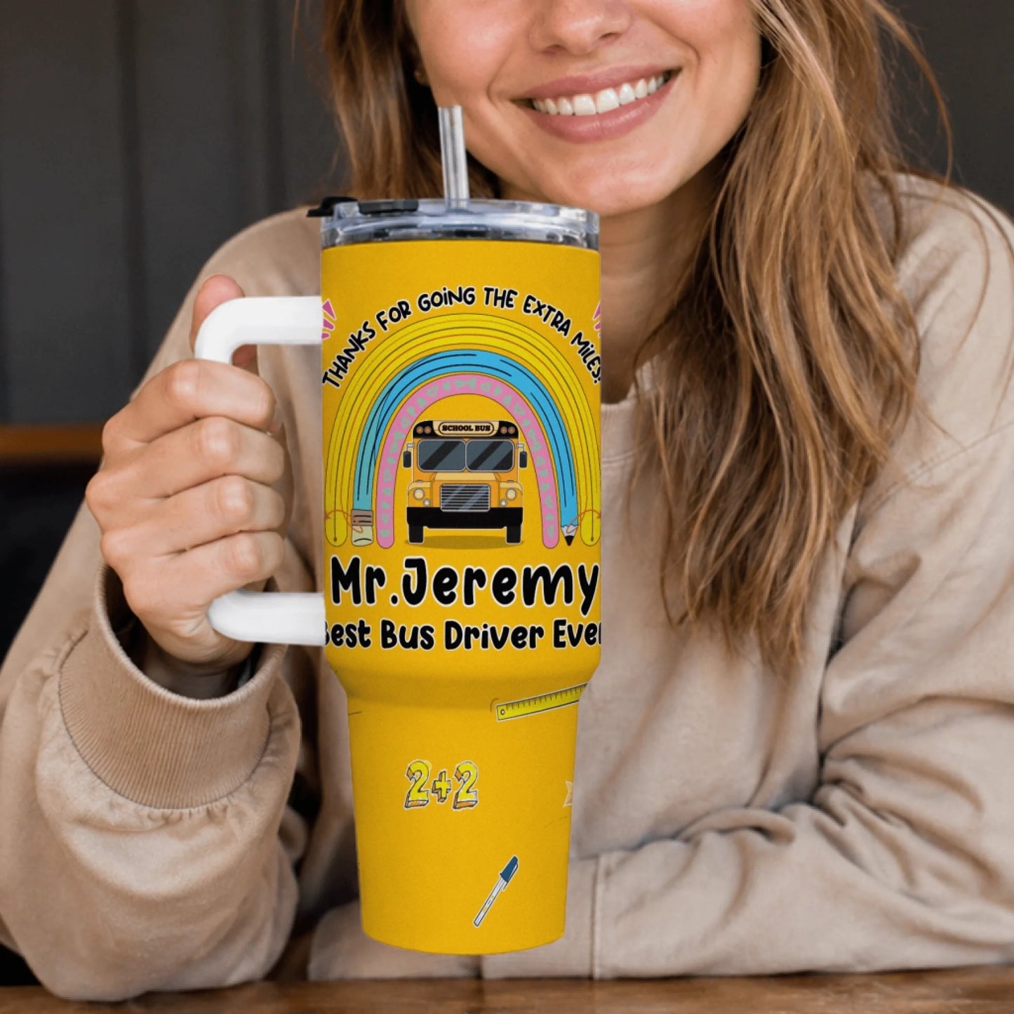 A smiling person holds a yellow handled travel tumbler decorated with a rainbow, school bus, and text "Mr. Jeremy Best Bus Driver Ever".