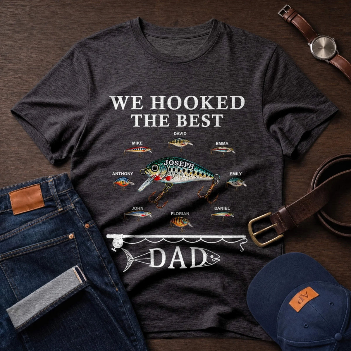 Dark gray t-shirt with fishing-themed design and text on a wooden surface with jeans, cap, and watch.