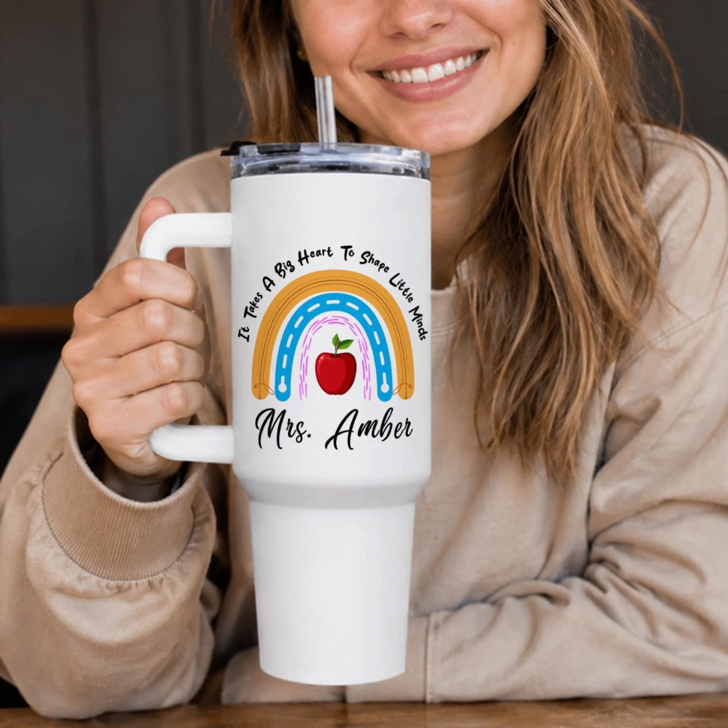 A smiling person holding a white handled tumbler with a rainbow, red apple, and 'Mrs. Amber' text.