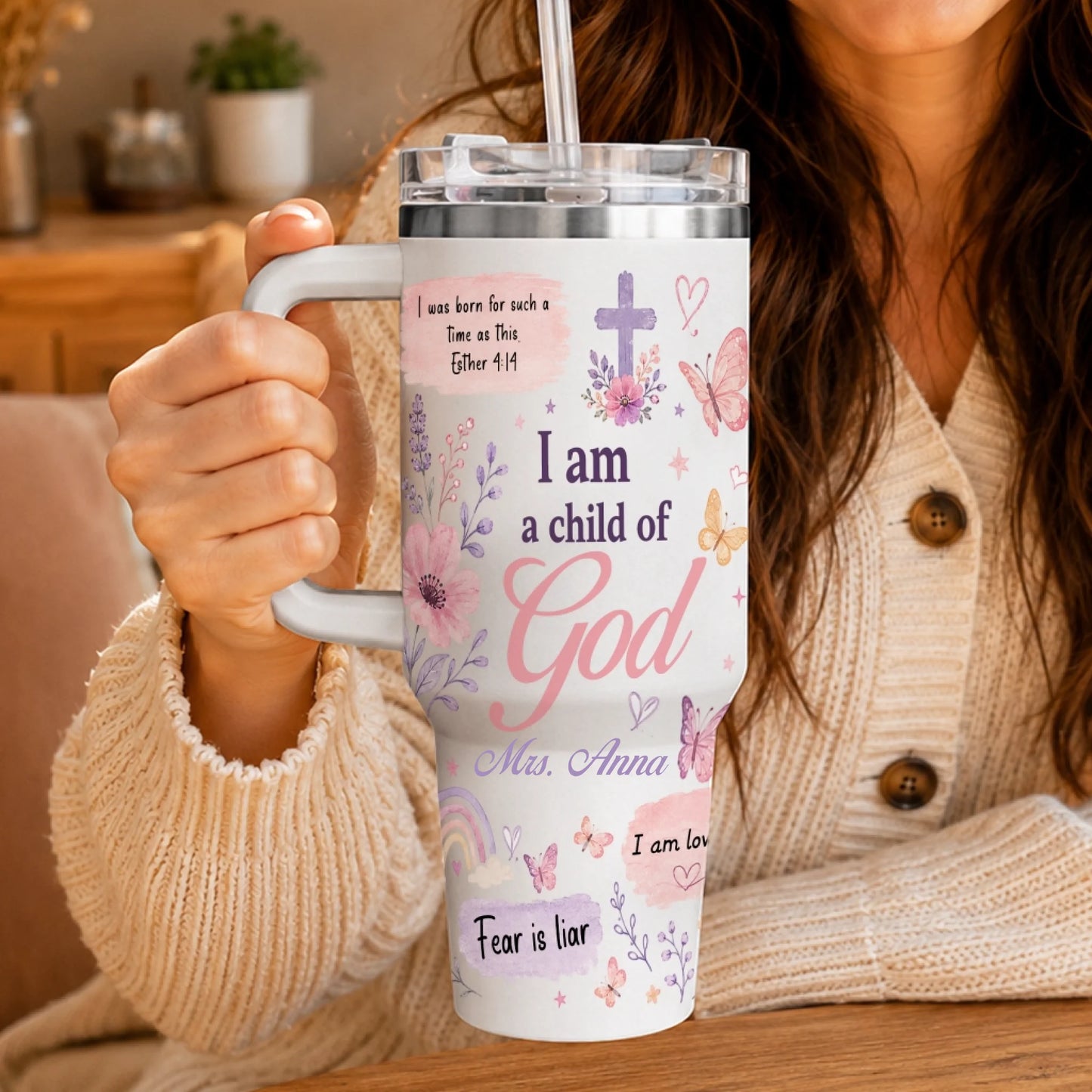 Close-up view of a Christian-themed tumbler held by a woman in a beige sweater, showing floral prints and scripture text.