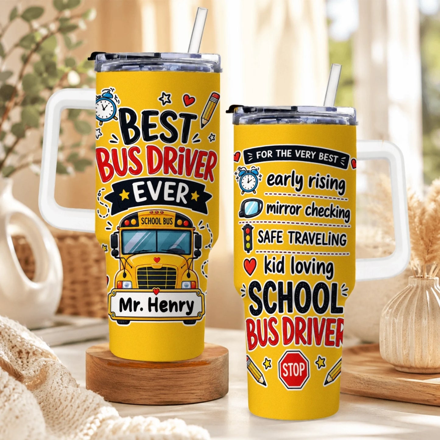 Two yellow tumblers with white handles and straws, featuring bus driver themed designs, sitting on a wooden surface.