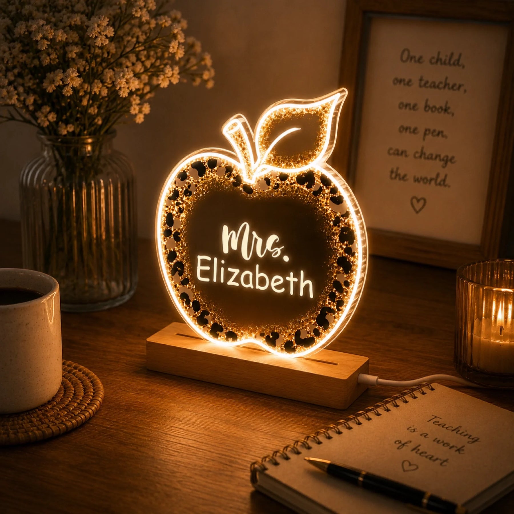 A glowing apple-shaped night light with a leopard print border and "Mrs. Elizabeth" text, on a desk with a mug, flowers, and a notebook.