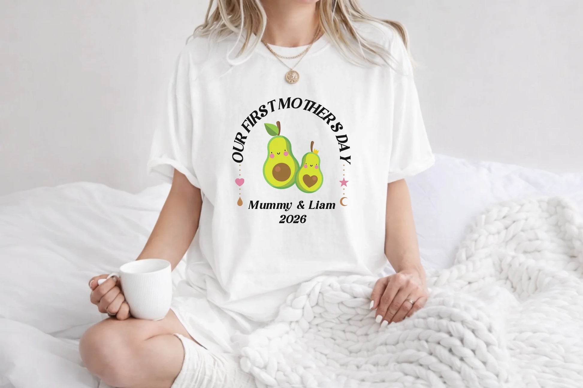 Person wearing a white t-shirt with 'Our First Mother's Day' text and avocado graphics, holding a mug.
