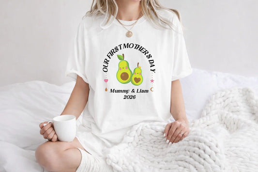 Person wearing a white t-shirt with 'Our First Mother's Day' text and avocado graphics, holding a mug.