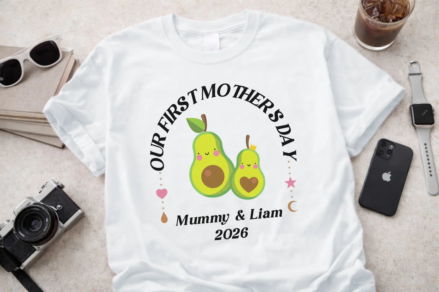 White t-shirt with 'Our First Mother's Day' design featuring two green pears, surrounded by small hearts and text, on a light surface with sunglasses, phone, and camera.