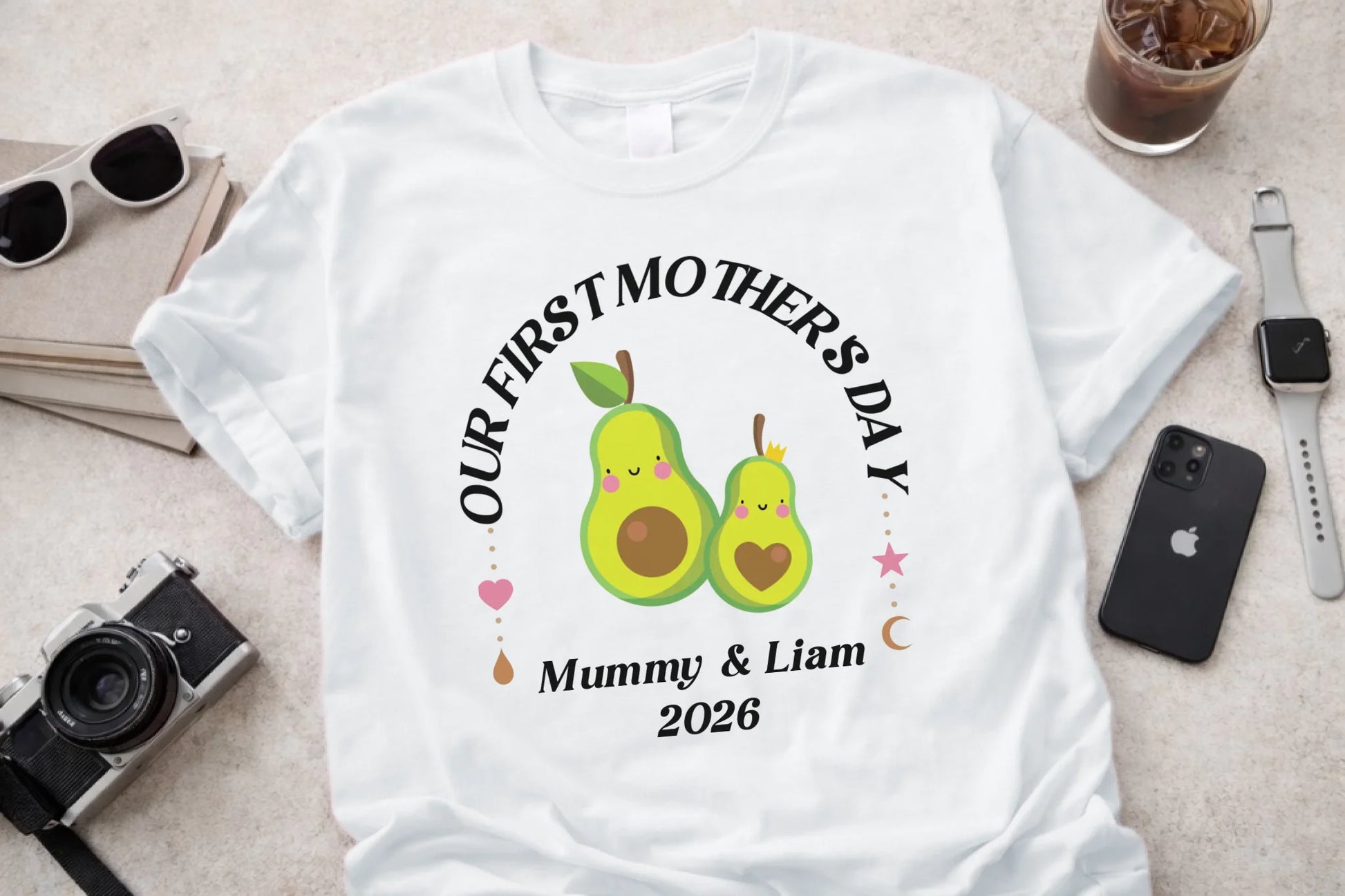 White t-shirt with 'Our First Mother's Day' design featuring two green pears, surrounded by small hearts and text, on a light surface with sunglasses, phone, and camera.