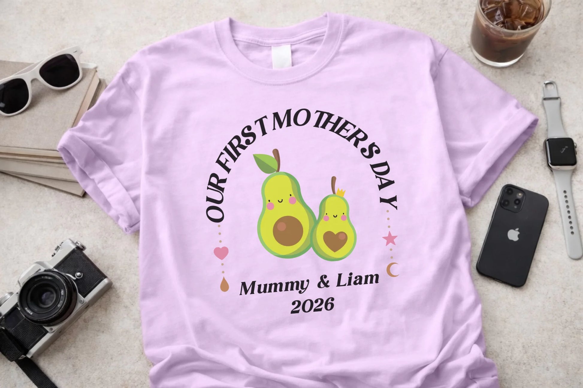 Pink t-shirt with 'Our First Mother's Day' text and pear graphics on a table with accessories.