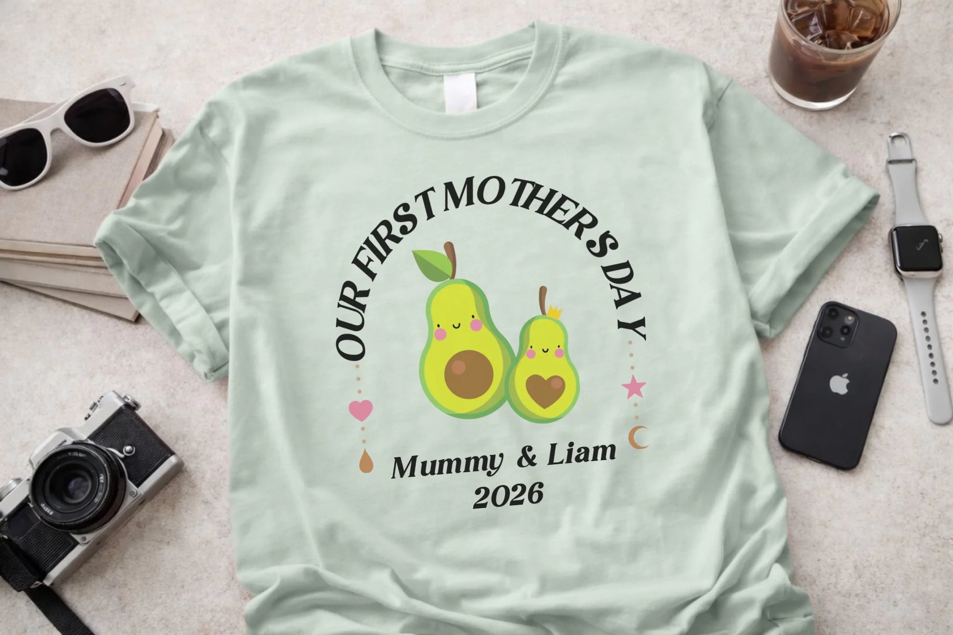Light green t-shirt with 'Our First Mother's Day' design featuring two cartoon pears, surrounded by sunglasses, a camera, a phone, and a watch on a light background.