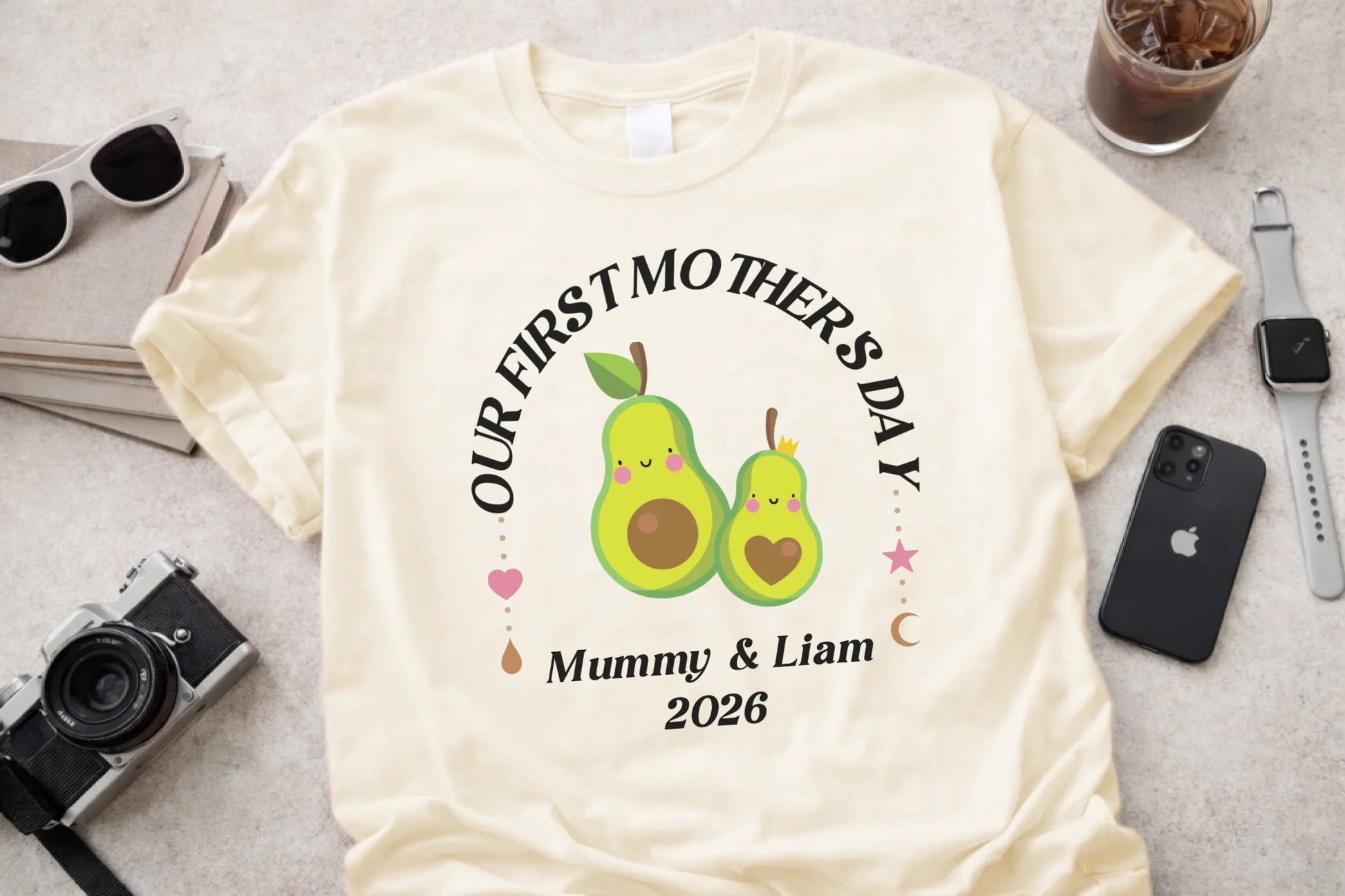 White t-shirt with 'Our First Mother's Day' design featuring two green pears, surrounded by a camera, sunglasses, and phone on a light gray surface.