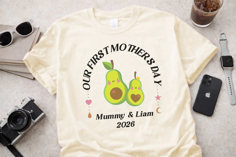 White t-shirt with 'Our First Mother's Day' design featuring two green pears, surrounded by a camera, sunglasses, and phone on a light gray surface.