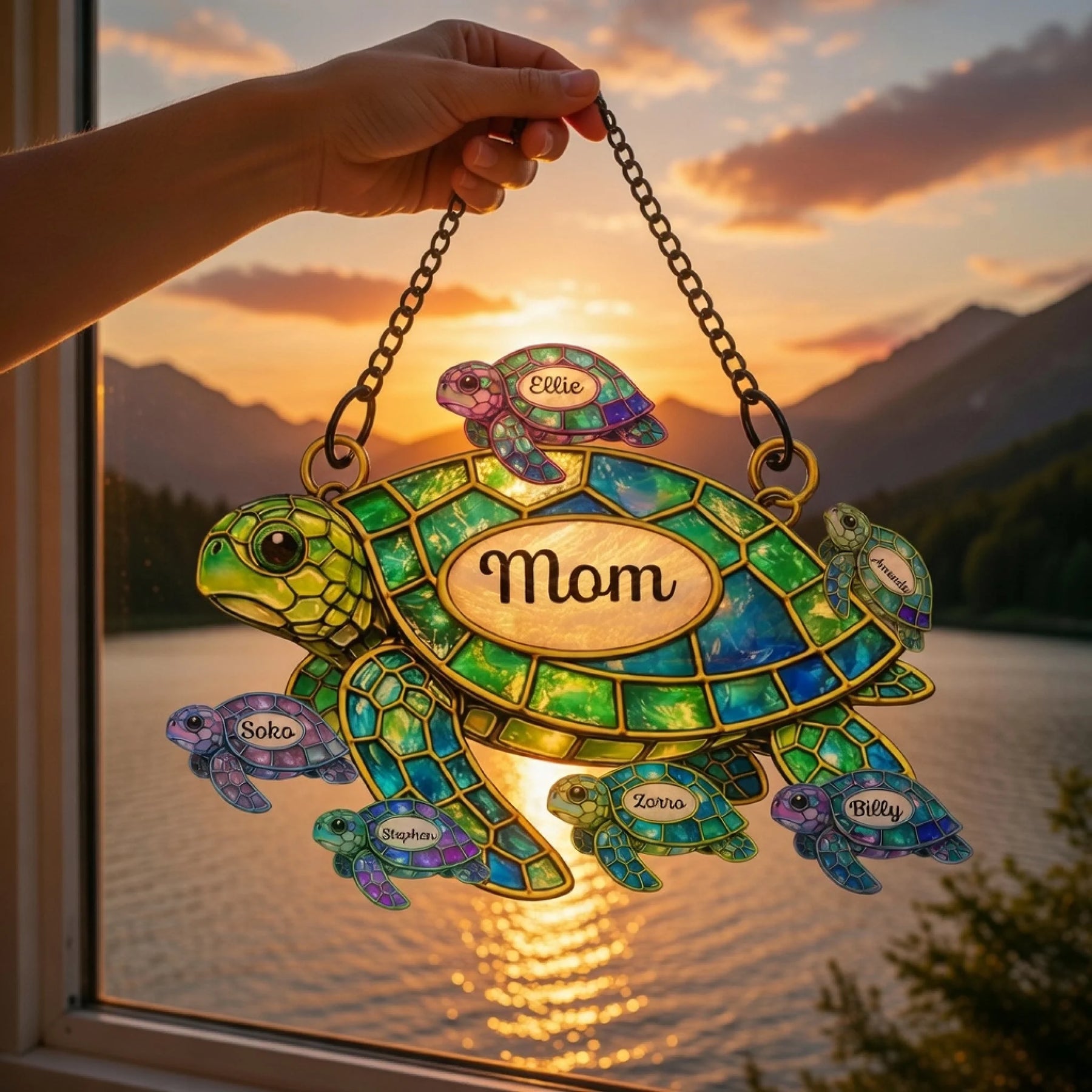 Decorative turtle ornament with 'Mom' on a chain, held by a hand against a sunset lake and mountain backdrop.