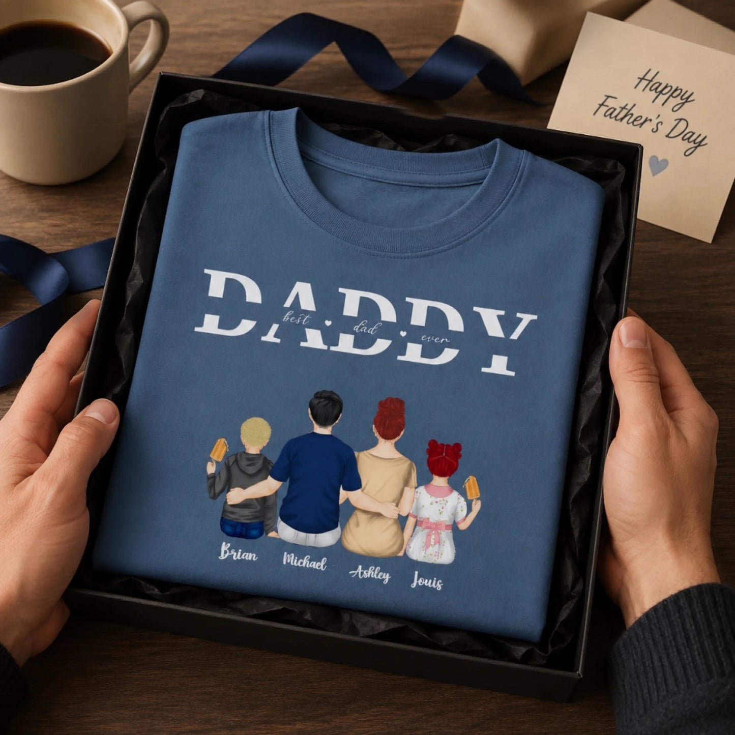 Blue t-shirt with family illustration and 'DADDY' text, held in hands on a wooden surface with a coffee cup and card.
