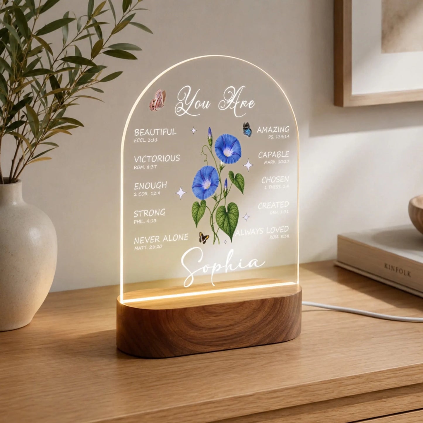 An illuminated arched acrylic night light with a wooden base, featuring "You Are" text, blue morning glory flowers, butterflies, and "Sophia." It is on a wooden dresser with a plant.