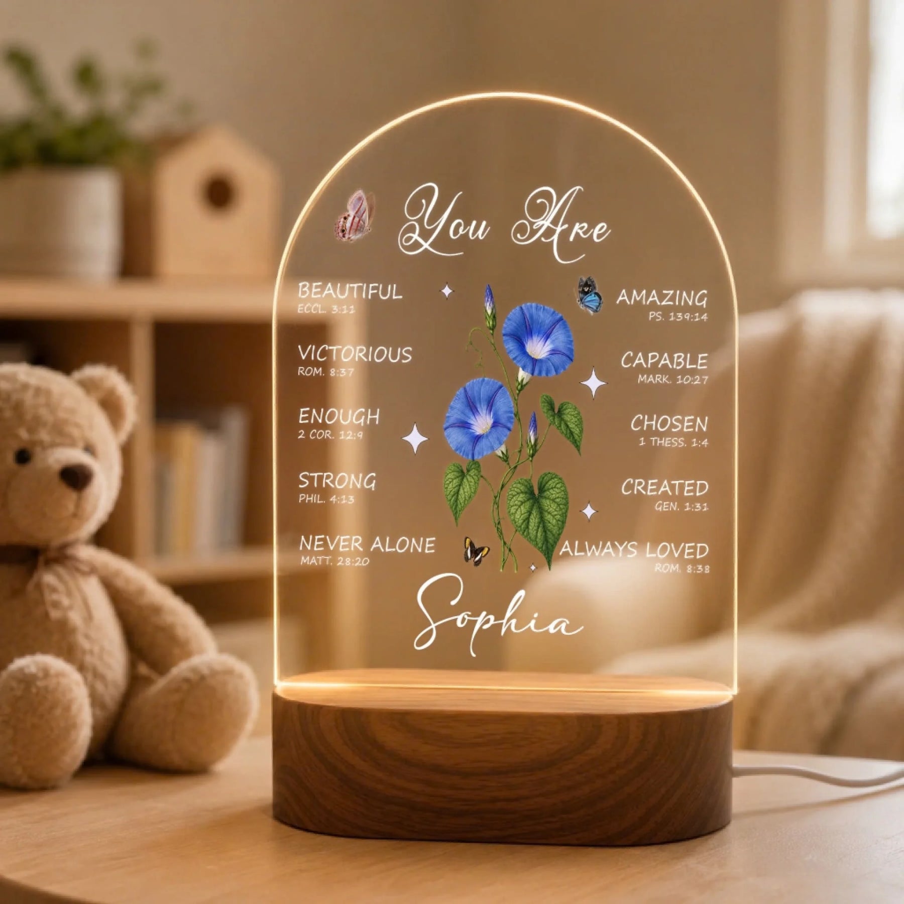An illuminated arched acrylic night light with a wooden base, displaying text, blue morning glory flowers, butterflies, and "Sophia." It is next to a brown teddy bear.