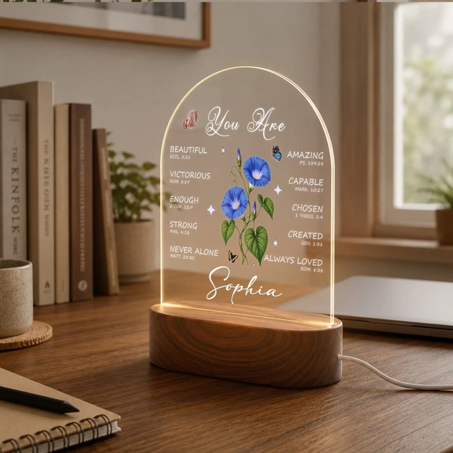 An illuminated arched acrylic night light with a wooden base, showing text, blue morning glory flowers, butterflies, and "Sophia." It is on a desk with books and a laptop.
