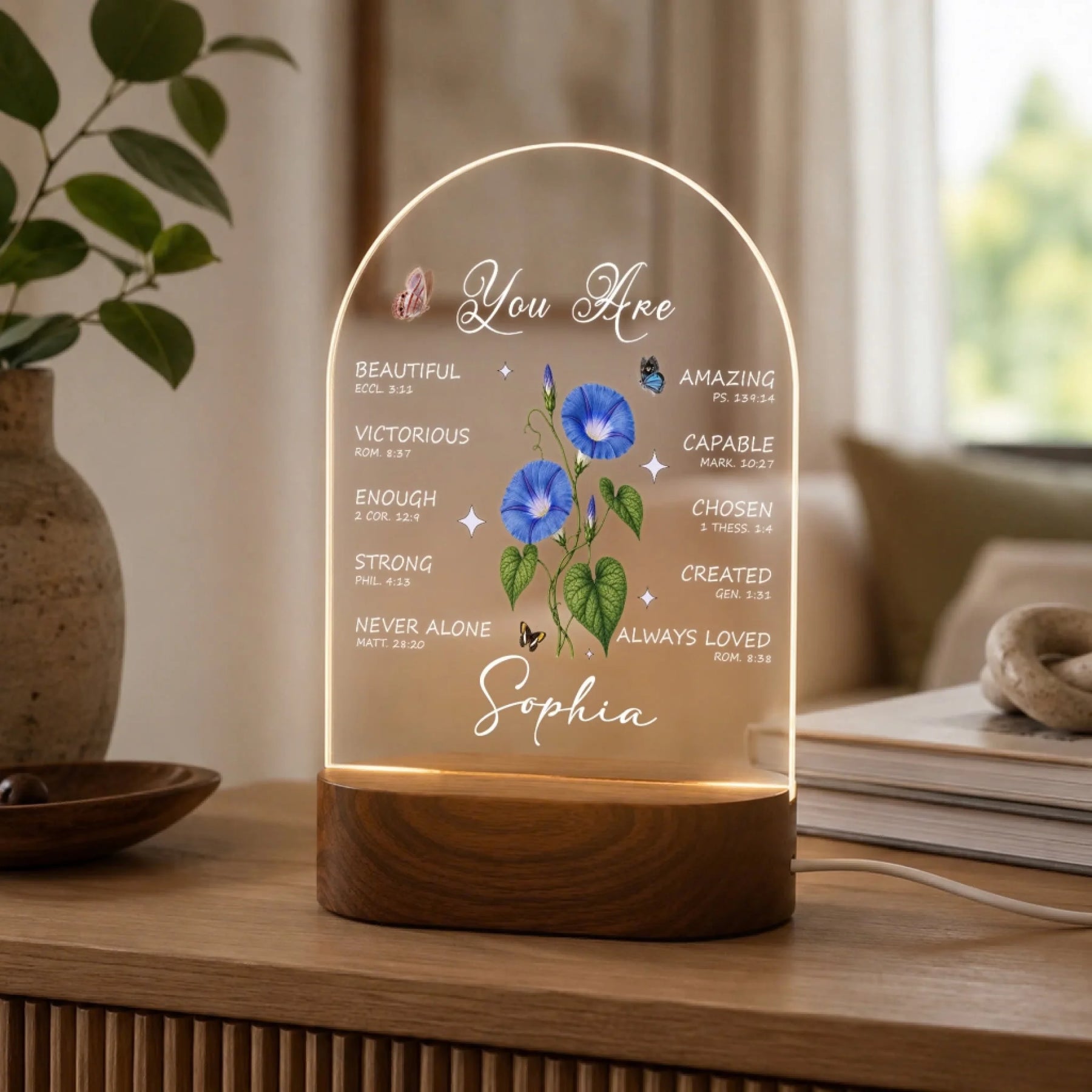 An illuminated arched acrylic night light with a wooden base, displaying text, blue morning glory flowers, butterflies, and "Sophia," in a bright home setting.