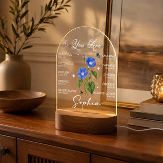 An illuminated arched acrylic plaque features blue morning glory flowers, butterflies, and empowering words like 'You Are Beautiful' and a visible name 'Sophia', on a wooden base, placed on a wooden cabinet with a plant and decor.