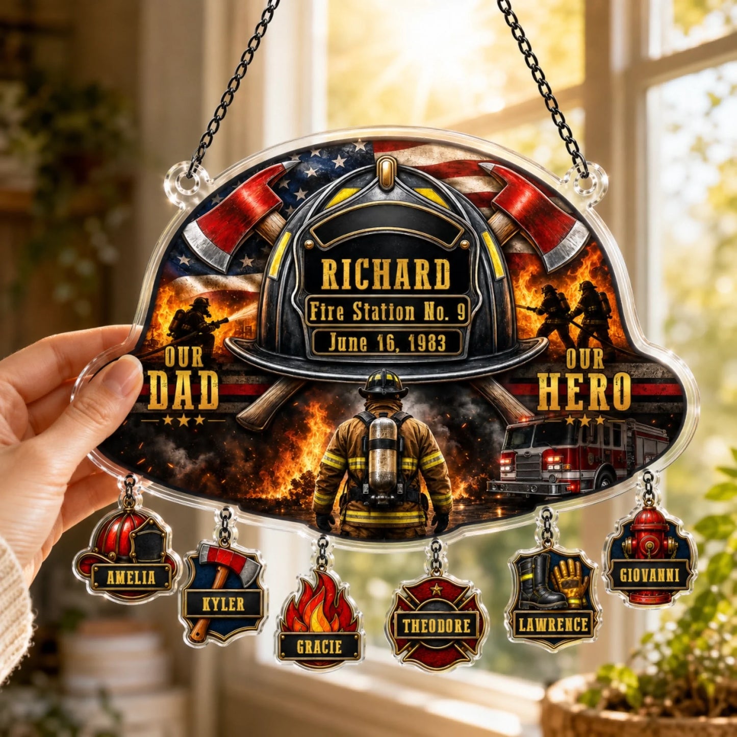 A hand holds a firefighter helmet-shaped suncatcher with a flag, helmet, axes, and text "OUR DAD", "OUR HERO". Six dangling charms below.