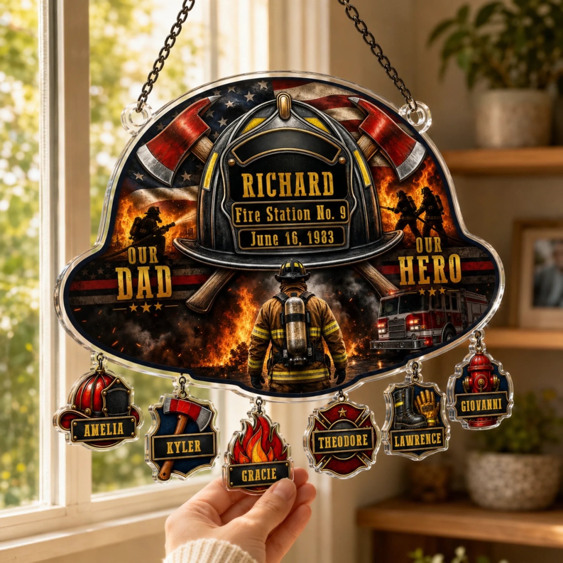 A hand holds a firefighter helmet-shaped suncatcher featuring a flag, helmet, axes, text, and six dangling personalized charms.