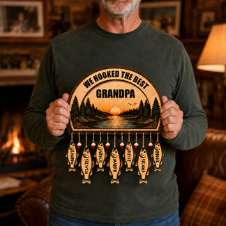 A person in a dark green shirt holds a wooden fishing-themed plaque. It reads "WE HOOKED THE BEST GRANDPA" with a sunset lake scene and seven fish-shaped name tags.