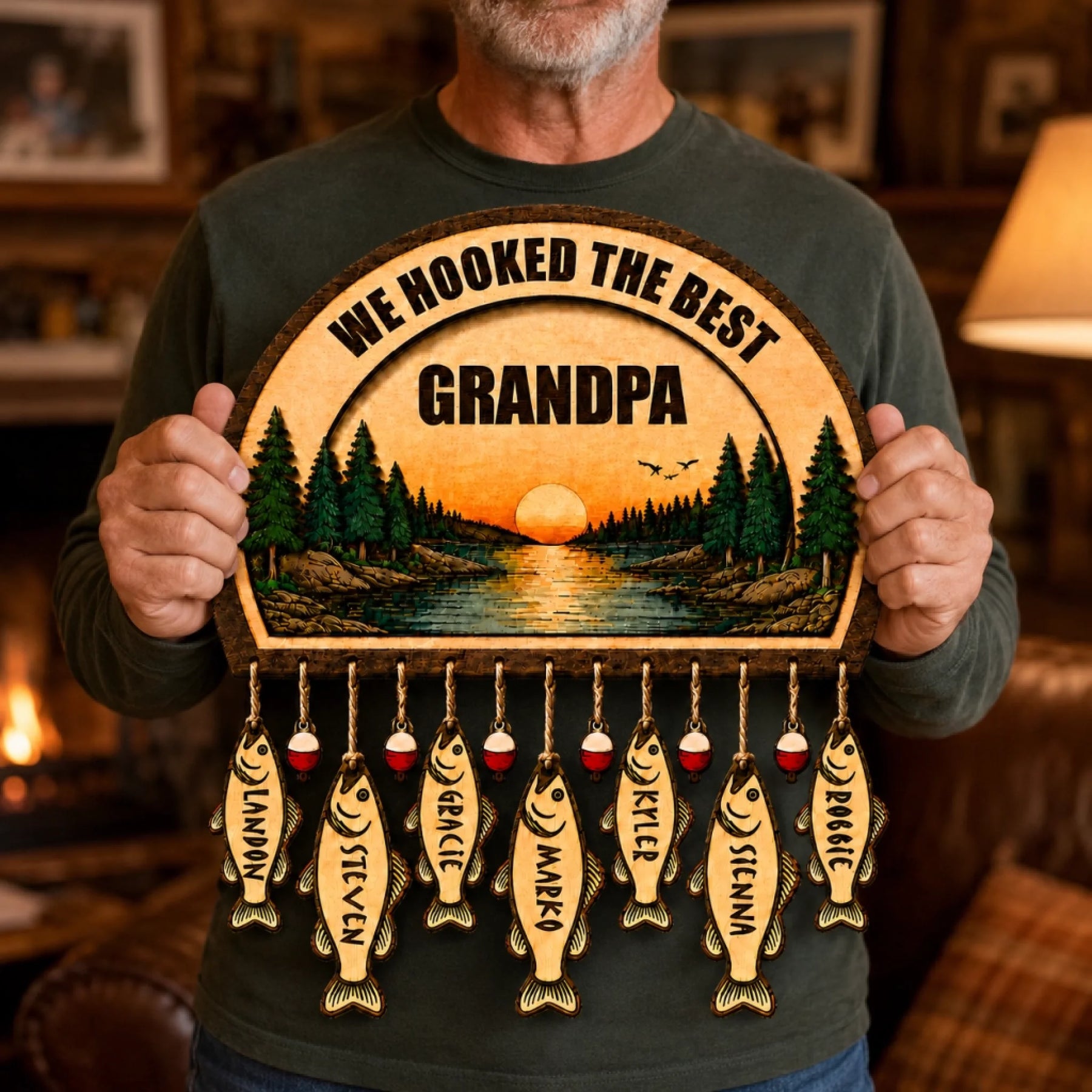 A close-up of a person holding a wooden fishing-themed plaque. It features "WE HOOKED THE BEST GRANDPA," a sunset lake scene, and seven fish-shaped tags with names.