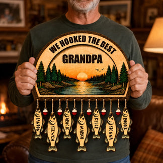 A close-up of a person holding a wooden fishing-themed plaque. It features "WE HOOKED THE BEST GRANDPA," a sunset lake scene, and seven fish-shaped tags with names.