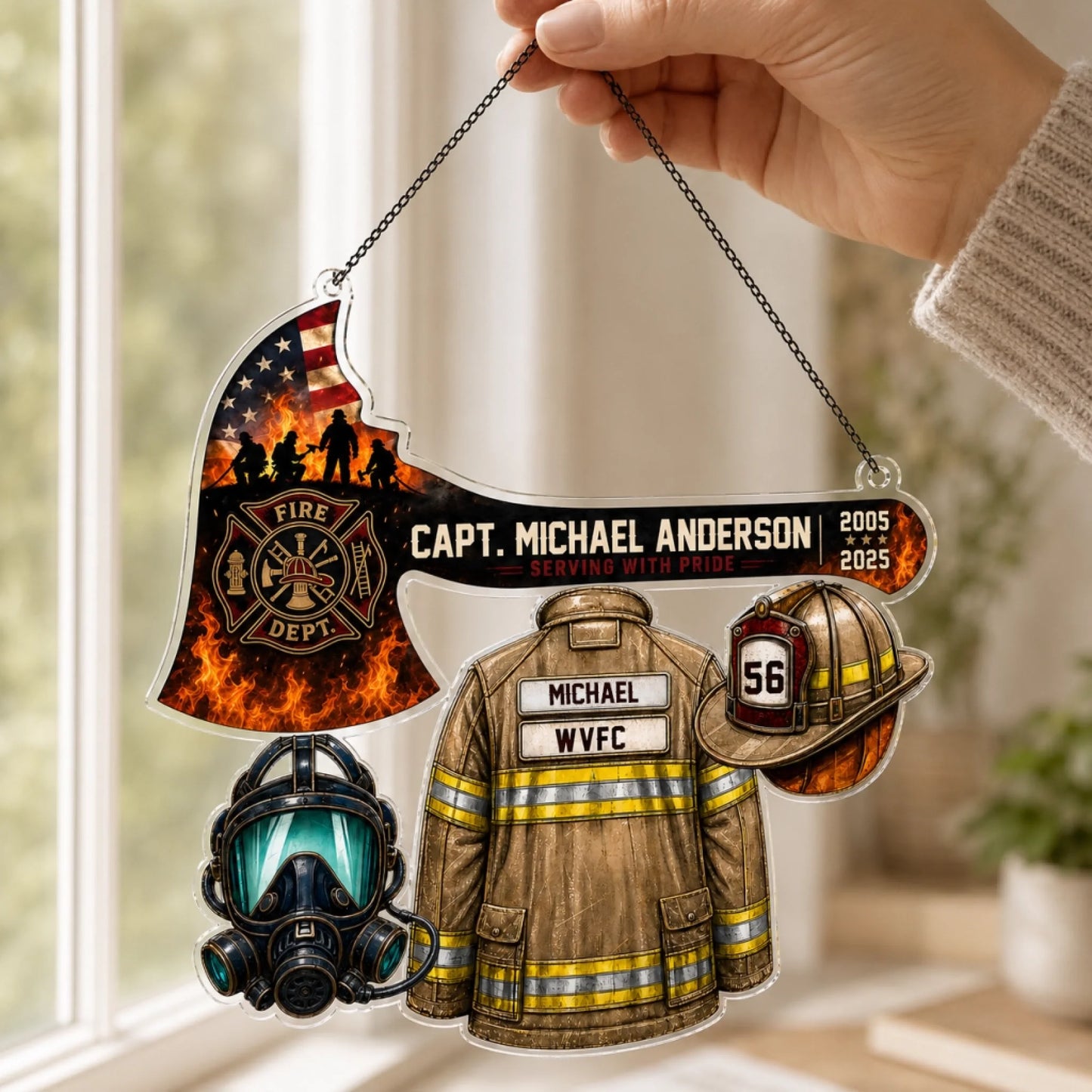 A hand holds an acrylic hanging firefighter ornament shaped like an axe with an American flag, a jacket, helmet, and mask.