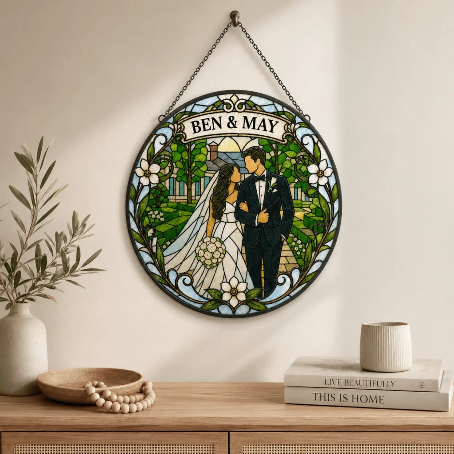 A circular stained glass panel depicting a bride and groom walking in a garden. The names 'BEN & MAY' are in a banner at the top. It hangs above a wooden console table with decorative items.