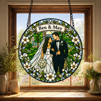 A circular stained glass piece depicting a bride and groom surrounded by greenery and a house, with a floral border. The names Ben & May are displayed at the top. It hangs in a wooden window frame with plants on the sill.