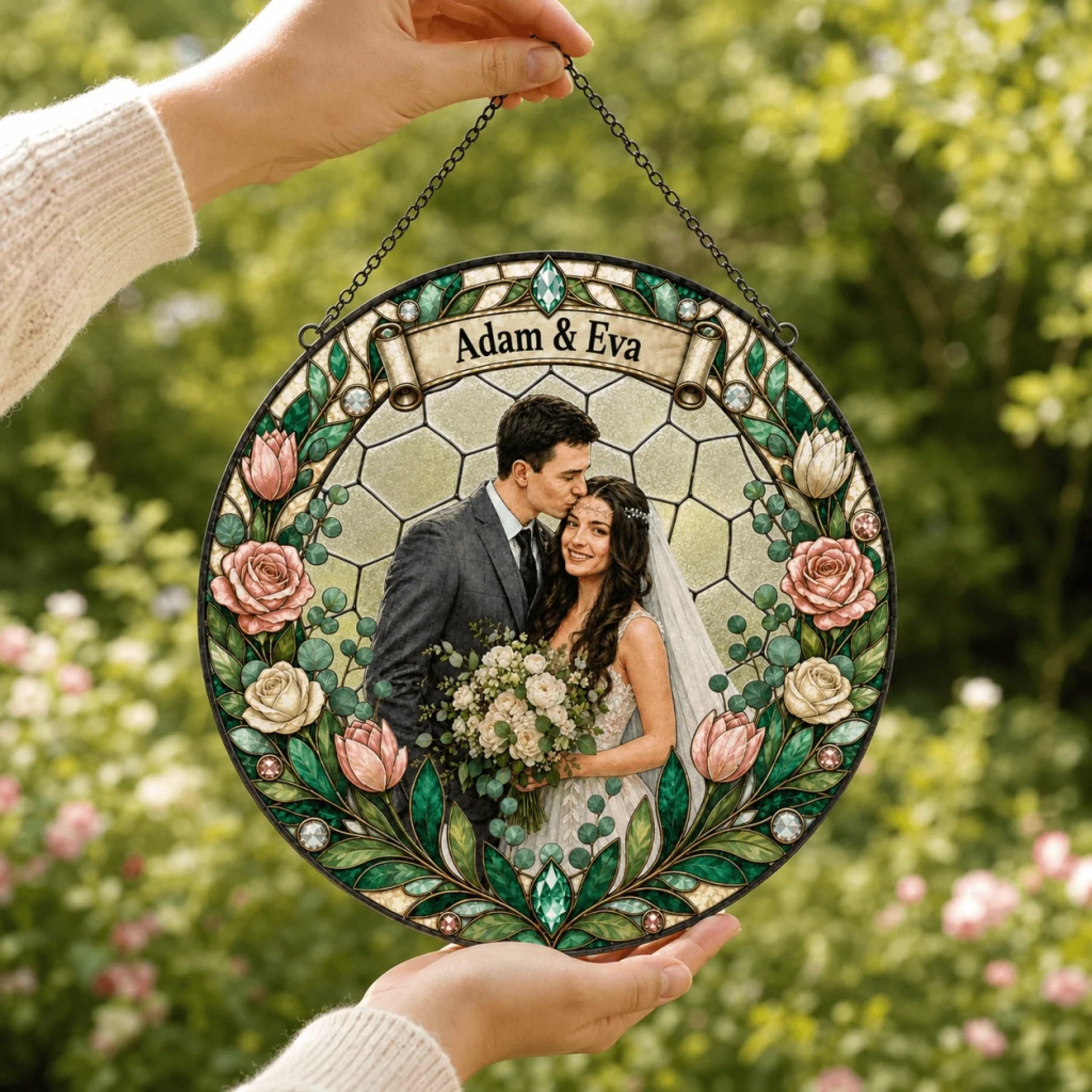 Hands hold a circular stained-glass style ornament, featuring a couple's photo and floral design, against a blurred green background.