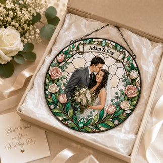 A circular stained-glass style ornament is nestled in an open gift box, surrounded by a white rose, ribbon, and a greeting card.