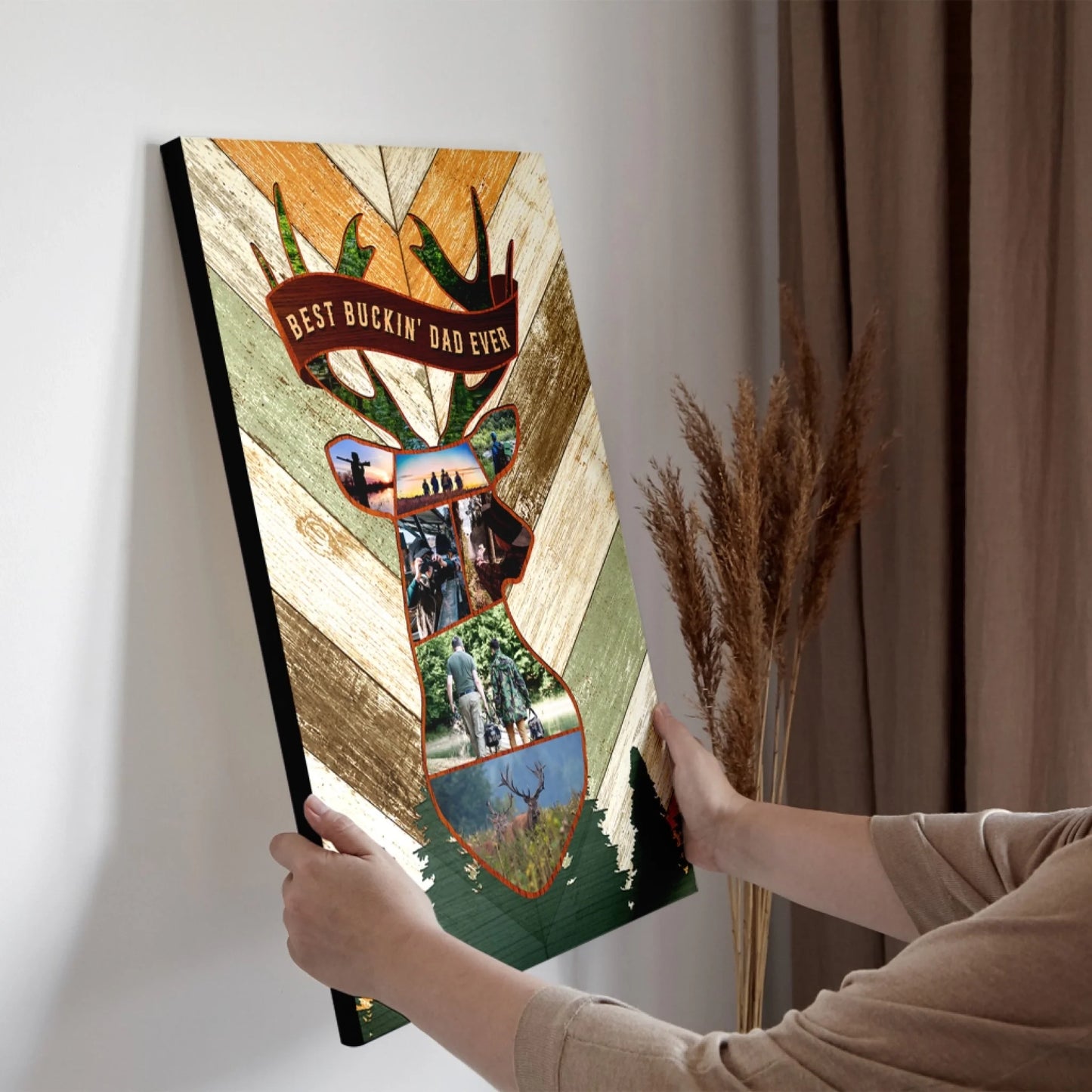 Two hands hold a vertical canvas print featuring a deer head collage with hunting photos and "BEST BUCKIN' DAD EVER" text, against a wall.