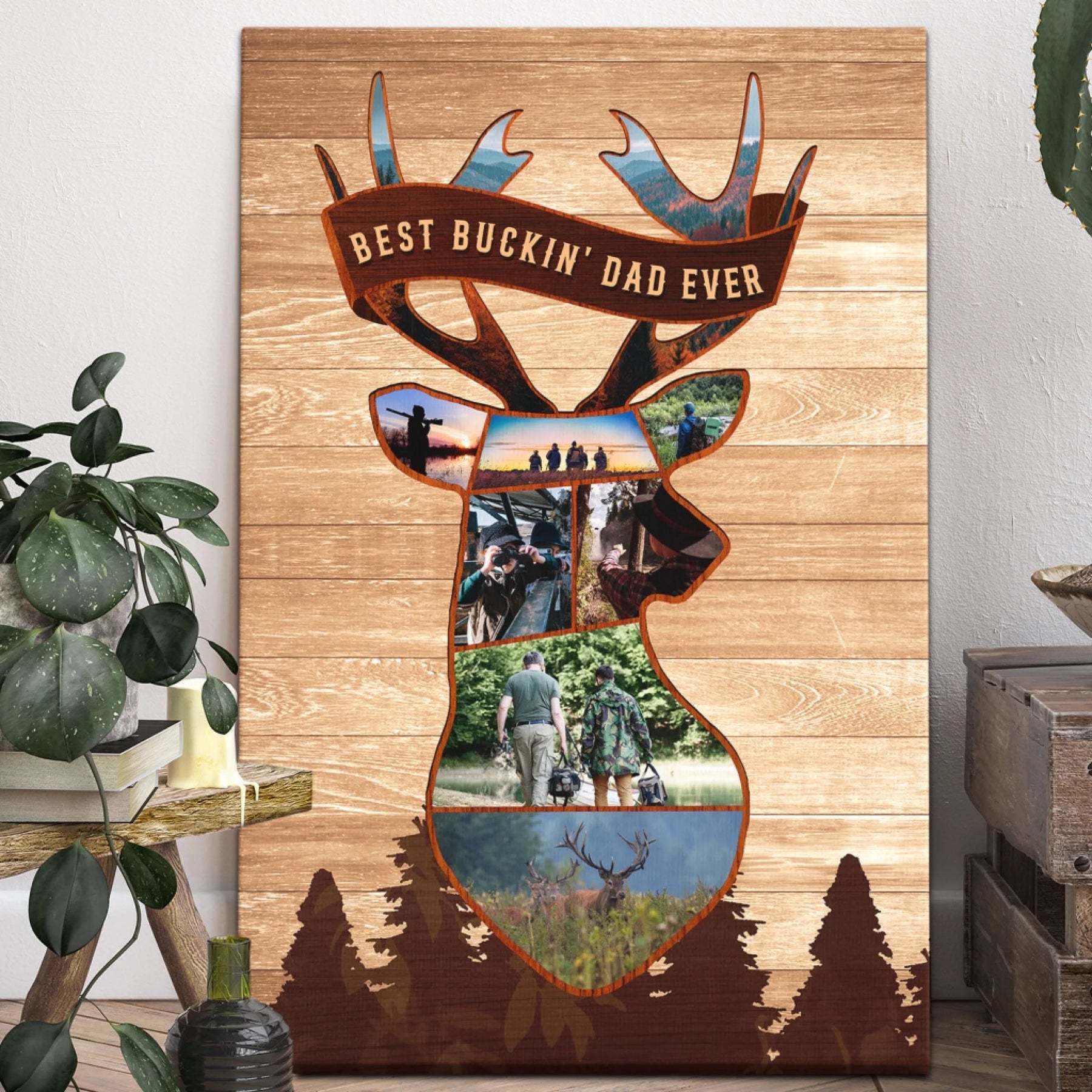 Vertical canvas print featuring a deer head photo collage, a banner reading "BEST BUCKIN' DAD EVER", and dark pine trees, displayed next to a houseplant and wooden crates.