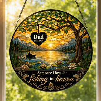 Round stained glass-style panel showing a sunset lake scene with a person fishing, mountains, trees, and water lilies. Text reads: "Dad 1950-2025" and "Someone I love is fishing in heaven."