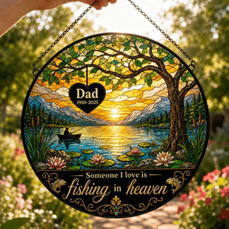 A hand holds a round, stained glass-style fishing scene panel outdoors. It depicts a sunset lake, a person fishing, and visible personalized text.