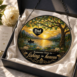 A round stained glass-style fishing scene panel with visible personalized text rests in an open gift box with tissue paper, beside a white flower and a card.