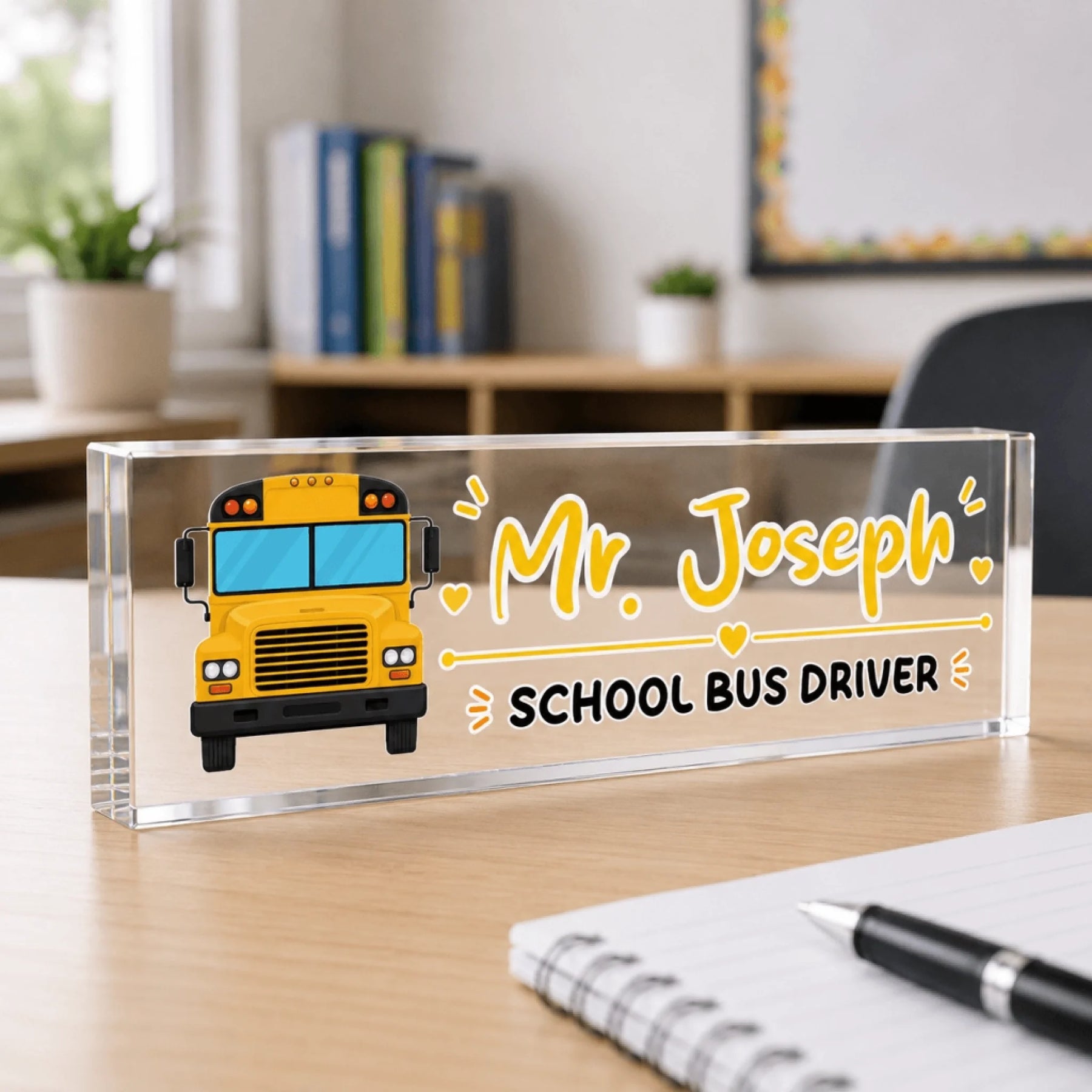 A clear acrylic desk sign with a cartoon yellow school bus and "Mr. Joseph" displayed on a desk with a notebook and pen.