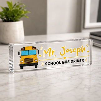 A clear acrylic desk sign for a school bus driver, showing a cartoon yellow bus and "Mr. Joseph", on a marble surface.