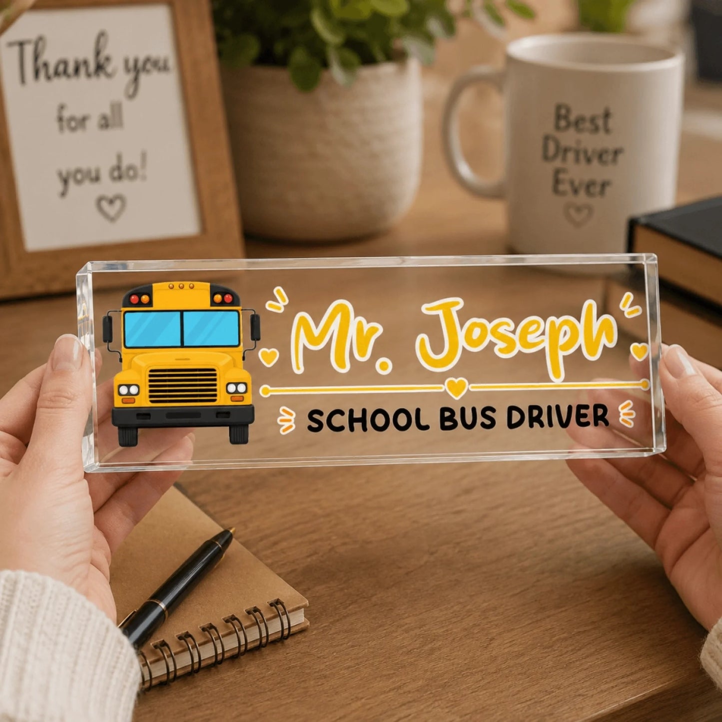 Two hands hold a clear acrylic desk sign with a yellow school bus illustration and the name "Mr. Joseph" on it.