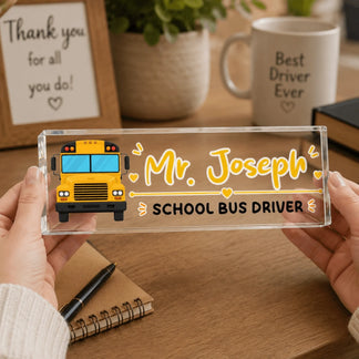 Two hands hold a clear acrylic desk sign with a yellow school bus illustration and the name "Mr. Joseph" on it.