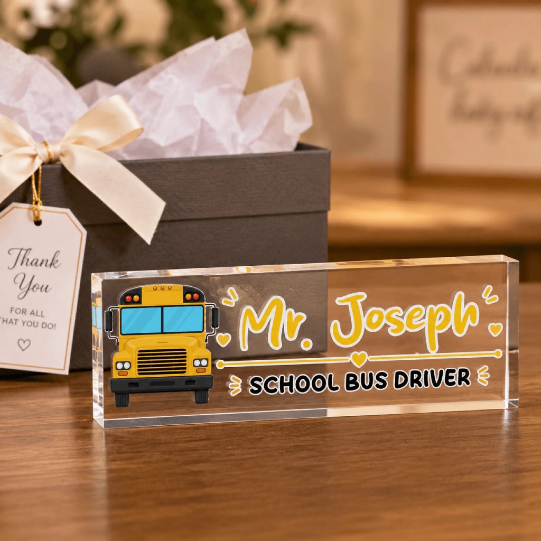 An acrylic desk sign for a school bus driver, featuring a yellow bus and "Mr. Joseph", next to a gift box with a tag.