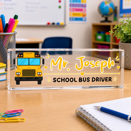 An acrylic desk sign for a school bus driver, featuring a yellow cartoon bus and the name "Mr. Joseph", on a classroom desk.