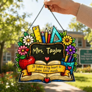 A hand holds up a colorful stained-glass style decorative plaque with "Mrs. Taylor" on its chalkboard, outdoors against foliage.