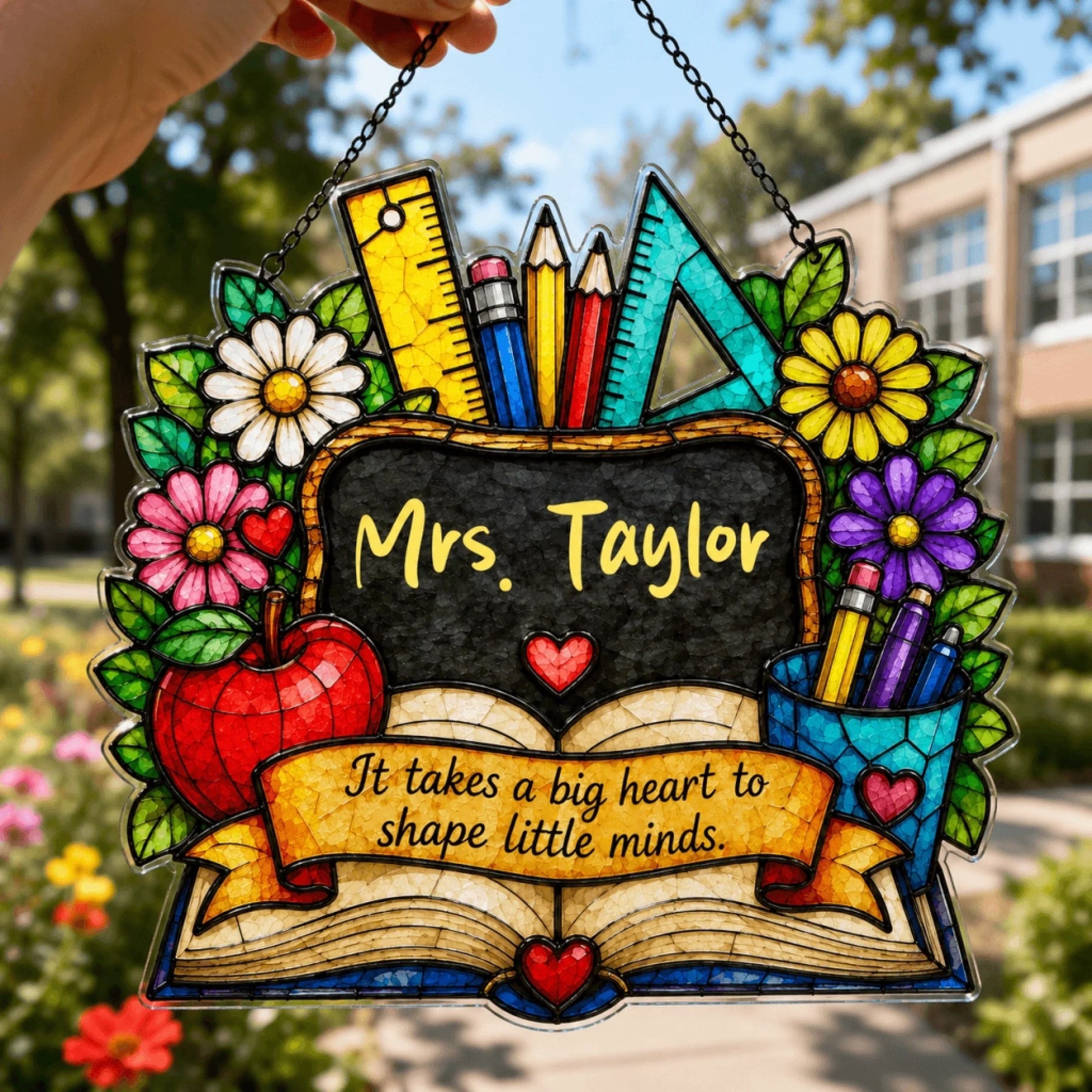 A colorful stained-glass style decorative plaque, featuring "Mrs. Taylor" on a chalkboard, an open book, and supplies, held outdoors.