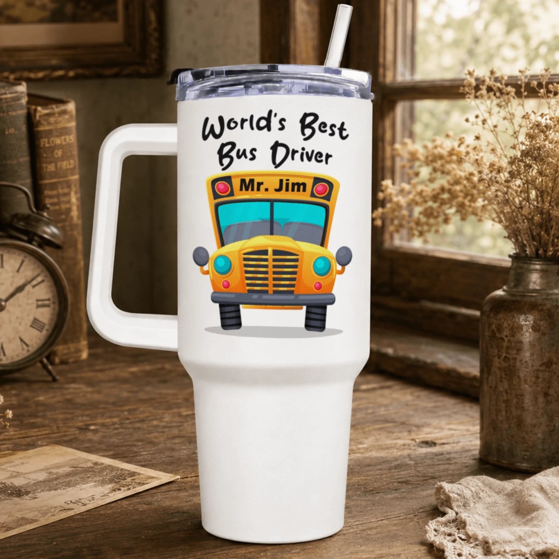 A white handled tumbler with a clear lid and straw features a cartoon yellow school bus and text "World's Best Bus Driver Mr. Jim", on a wooden desk.