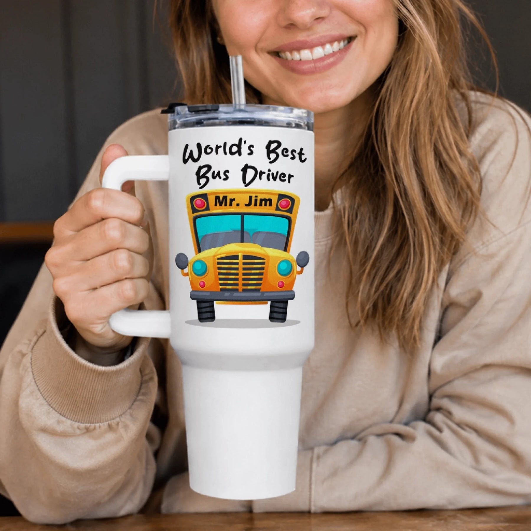 A smiling woman holds a large white handled tumbler with a straw. The tumbler features a cartoon school bus and text 'World's Best Bus Driver Mr. Jim'.