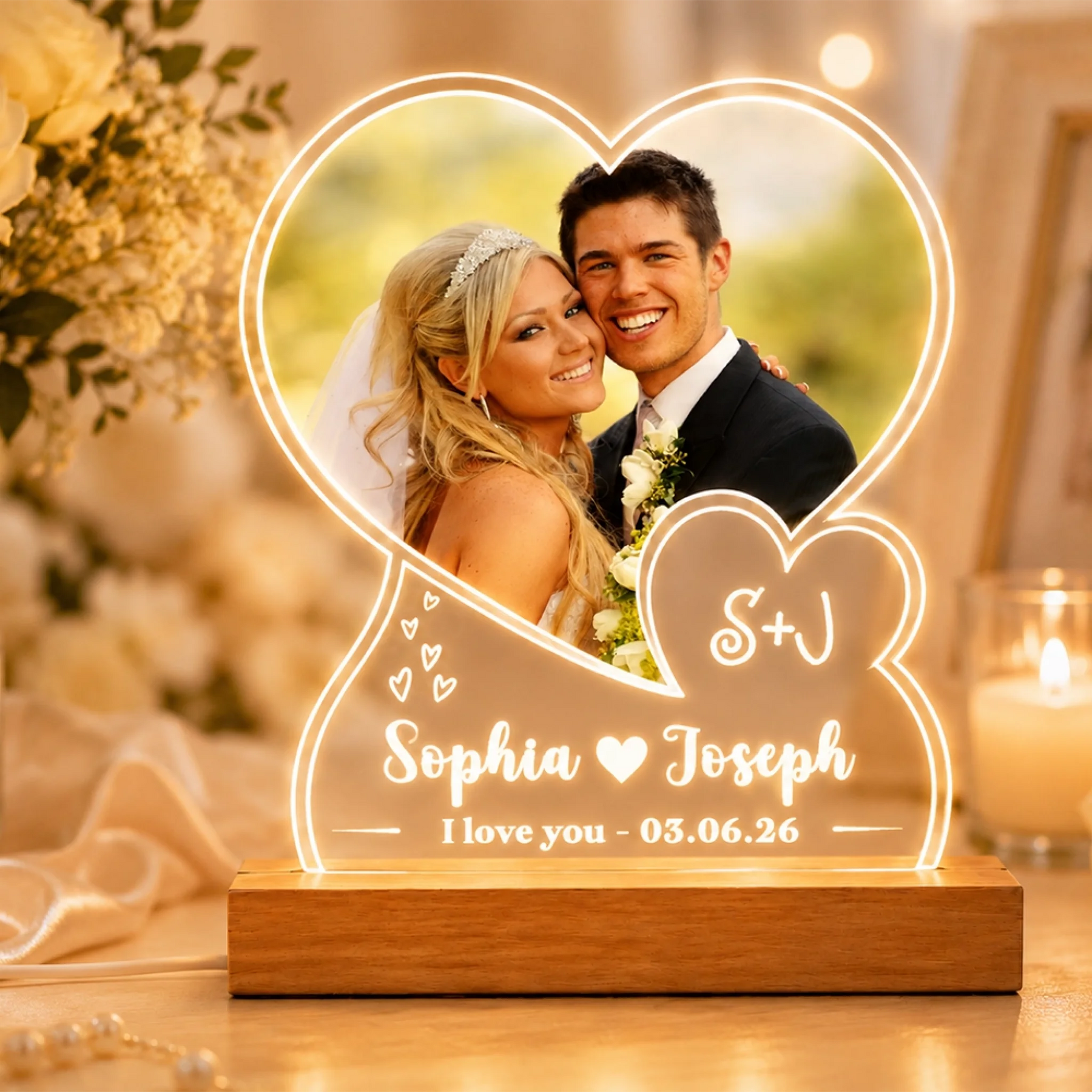 A heart-shaped illuminated custom light displaying a wedding photo of a couple, with "Sophia ❤️ Joseph" and "I love you - 03.06.26" inscribed on it, placed on a wooden base, surrounded by blurred flowers and a candle.