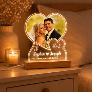 A heart-shaped custom photo light, featuring a wedding picture and personalized text, glows on a nightstand beside a lit candle and a vase of dried flowers in a dimly lit bedroom.