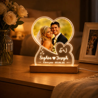A heart-shaped custom photo light, illuminated with a wedding image and engraved text, sits on a wooden desk with a vase of white roses and a framed photo in a warm room.